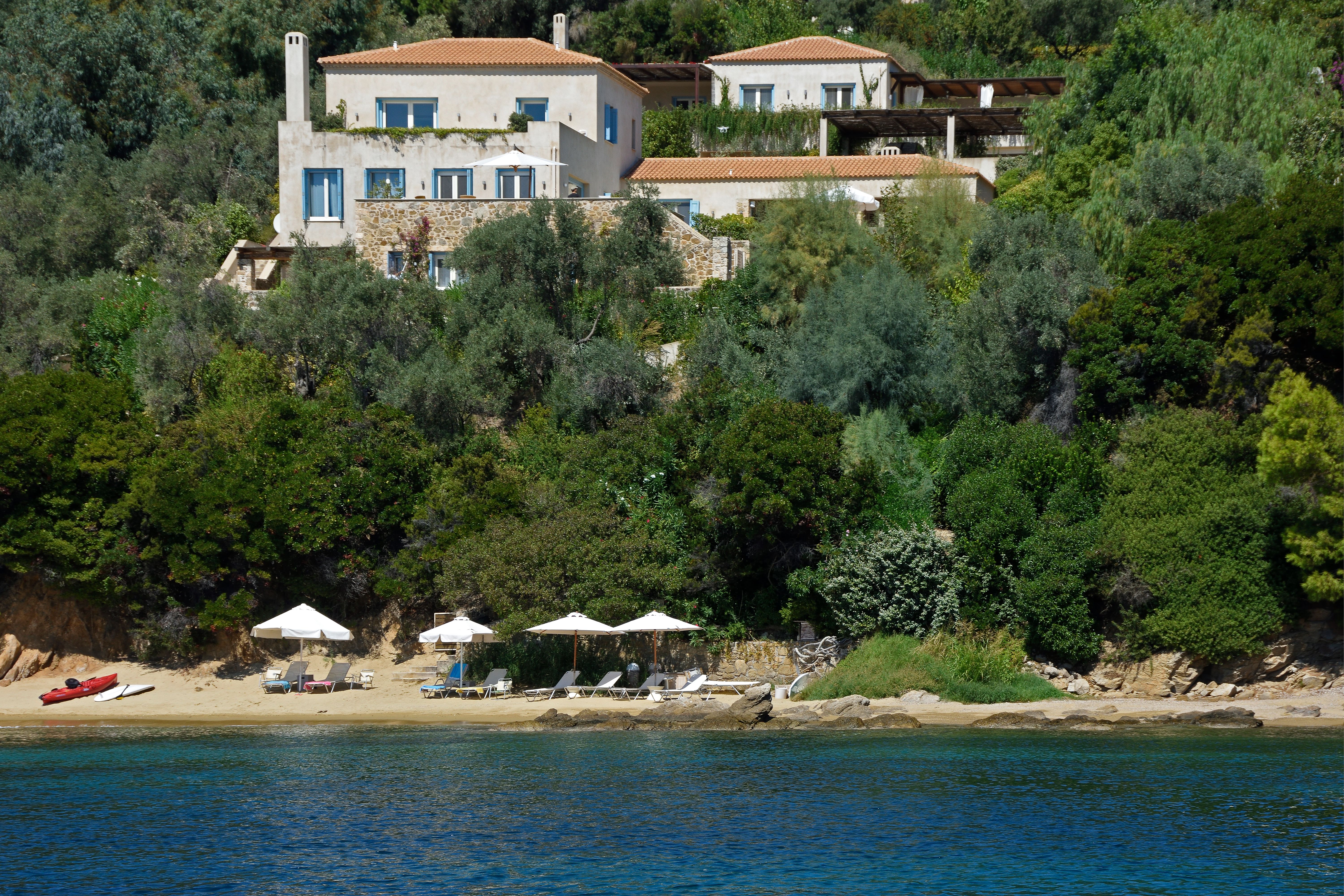 Villa Goldie above the trees, with sunbeds and umbrellas on the beach below, seen from the sea